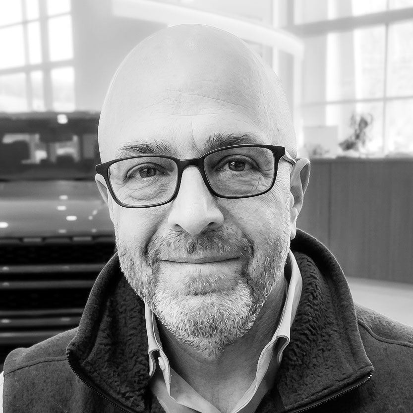 Portrait of a smiling bald man with black-framed glasses in a bright indoor lobby.