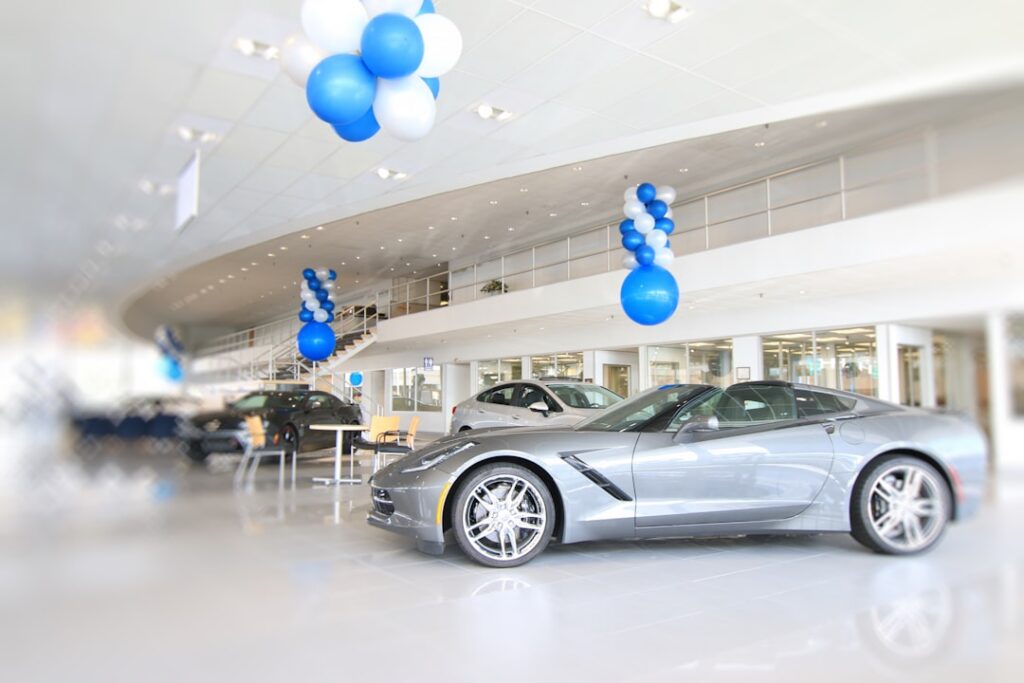 Showroom interior with a silver sports car on display and blue‑white balloons overhead. Other cars and seating are visible in the bright showroom.