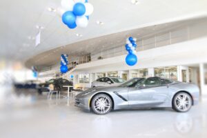 Showroom interior with a silver sports car on display and blue‑white balloons overhead. Other cars and seating are visible in the bright showroom.