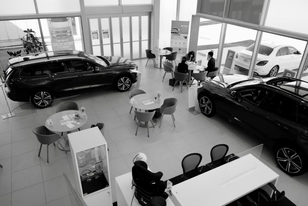 Interior of a car dealership showroom with two dark cars on display, round tables, and customers chatting near the windows.
