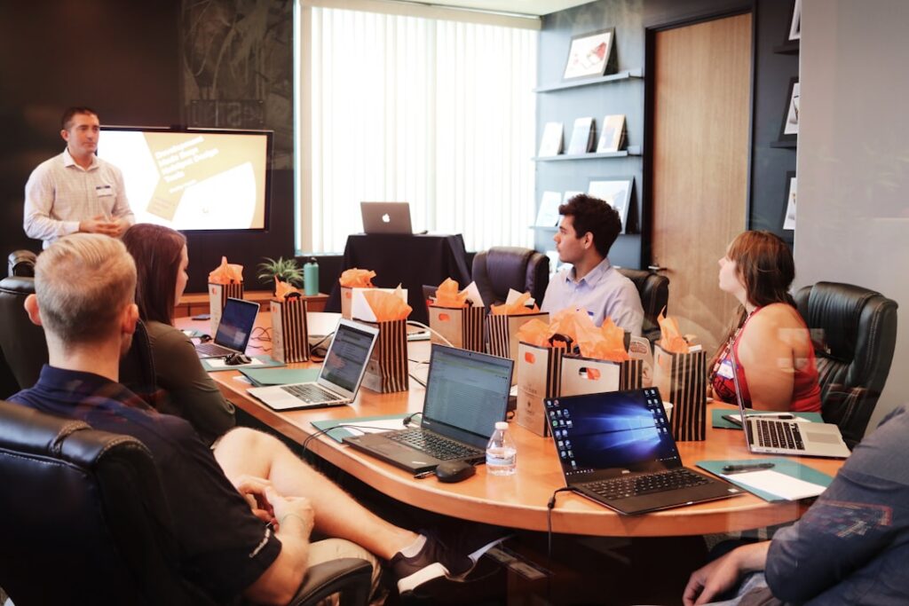 Presenter giving a talk to a round conference table of coworkers, laptops open and gift bags lined along the table in a meeting room