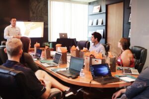 Dealership-sales-technology-best-practices - proactive training solutions Presenter giving a talk to a round conference table of coworkers, laptops open and gift bags lined along the table in a meeting room