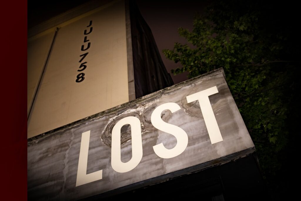 Weathered storefront sign reading lost, angled upward with a dark night scene and a tree in the background.