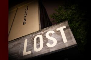 Weathered storefront sign reading lost, angled upward with a dark night scene and a tree in the background.