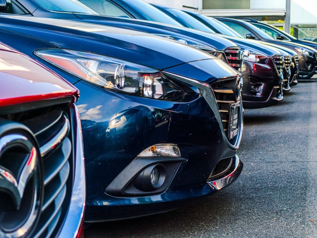 Row of modern cars parked side by side in a dealership lot, foreground blue car front and grille visible