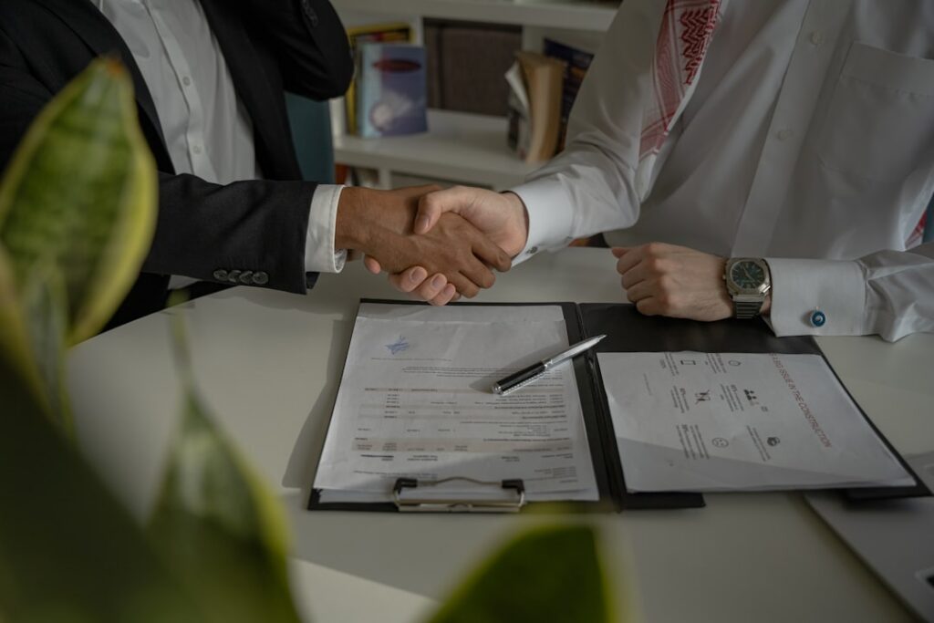 Two professionals shake hands over a desk with a signed documents binder, signaling an agreement or contract signing.