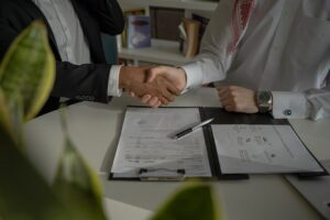 Used-car-sales-training-hold-gross - proactive training solutions Two professionals shake hands over a desk with a signed documents binder, signaling an agreement or contract signing.