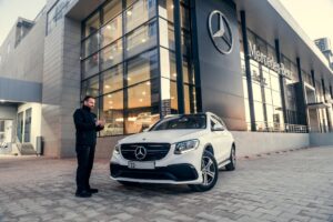 Man standing beside a white Mercedes SUV outside a Mercedes-Benz showroom, looking at his phone.